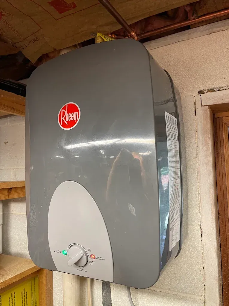 Gray Rheem wall-mounted water heater on an indoor wall, with a control dial near the bottom.