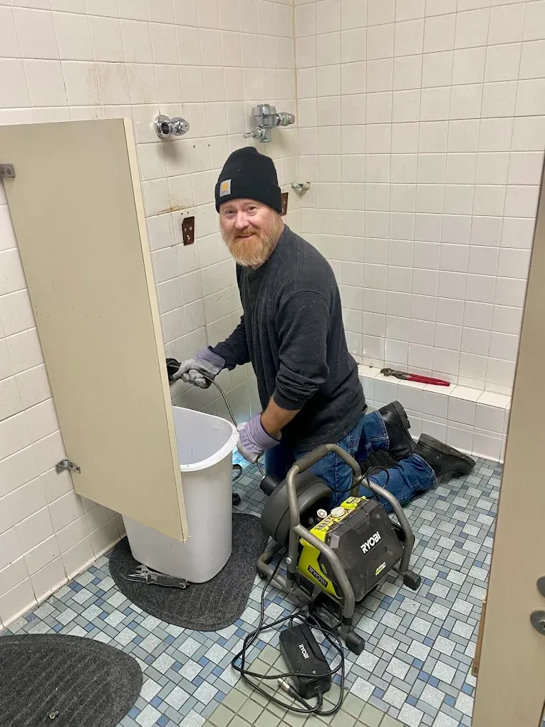 Worker using drain-cleaning equipment in a tiled restroom, cleaning a toilet drain.