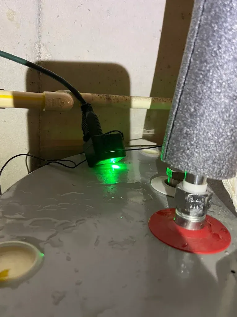 Green-lit device under a sink next to pipes and a red-capped fitting with water on the floor