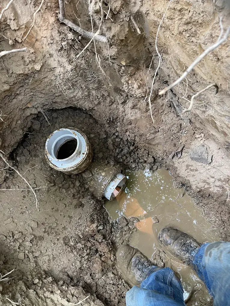 Open sewer pipe in muddy trench with standing water and a worker’s boots beside it