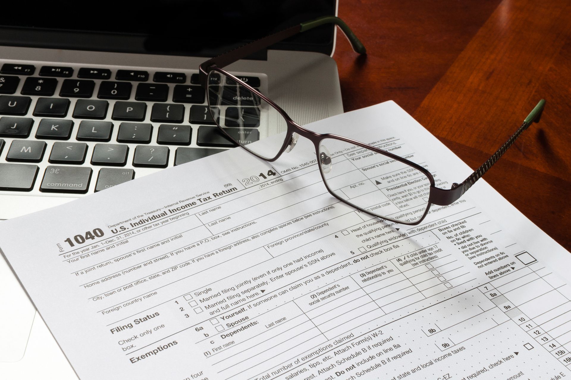 Laptop, tax form, and eyeglasses on a wooden surface, suggesting tax preparation or financial work.