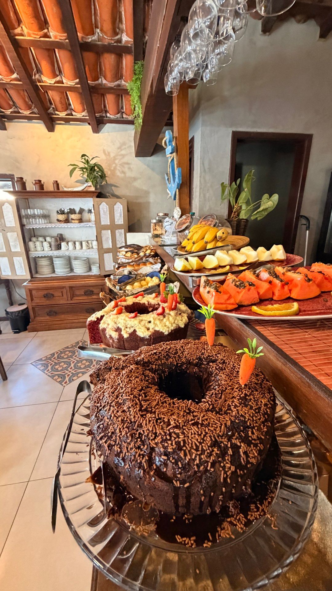 Bolo de chocolate numa mesa de buffet com travessas de frutas, num ambiente rústico interior