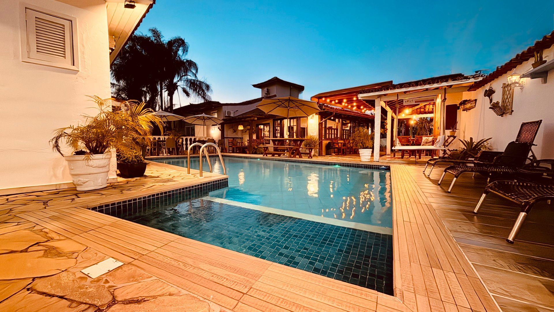 Pátio de resort junto à piscina ao pôr do sol, com água azul-turquesa, espreguiçadeiras e luzes aconchegantes