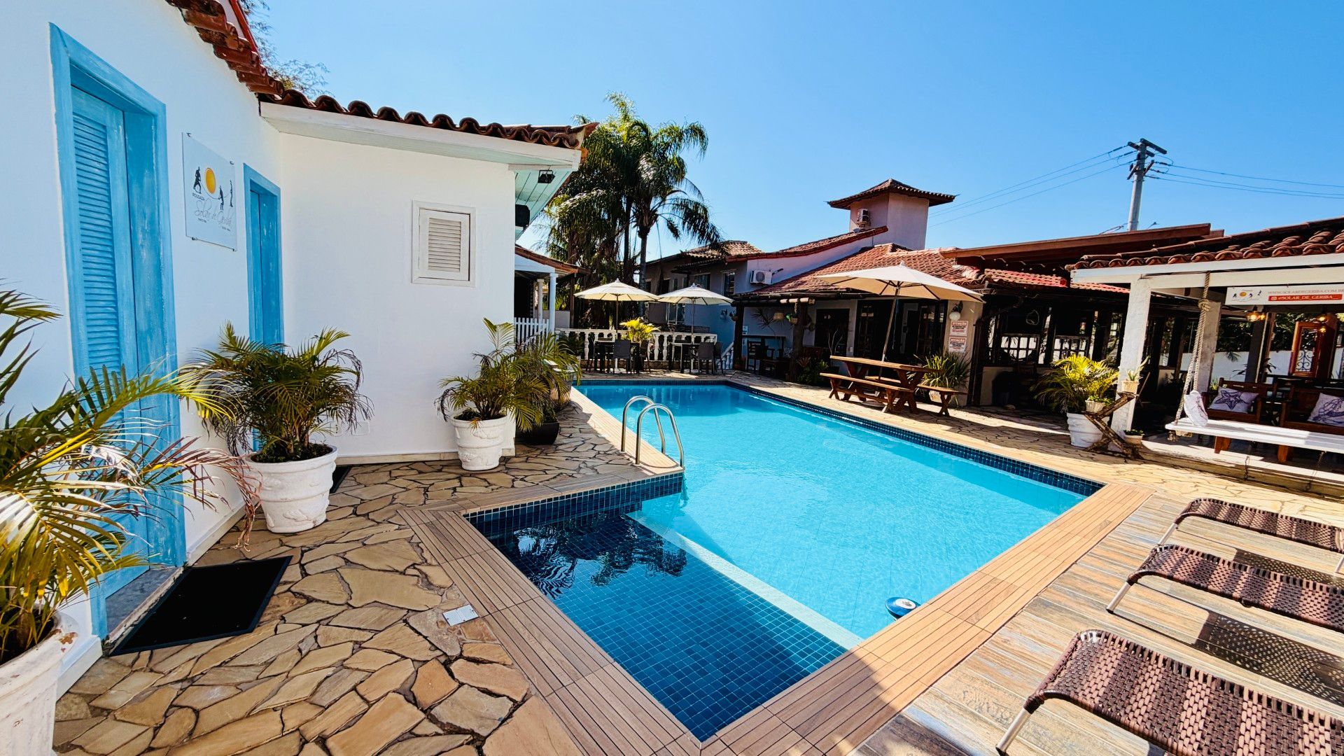 Pátio ensolarado com uma piscina azul ao lado de edifícios brancos e azuis e espreguiçadeiras.