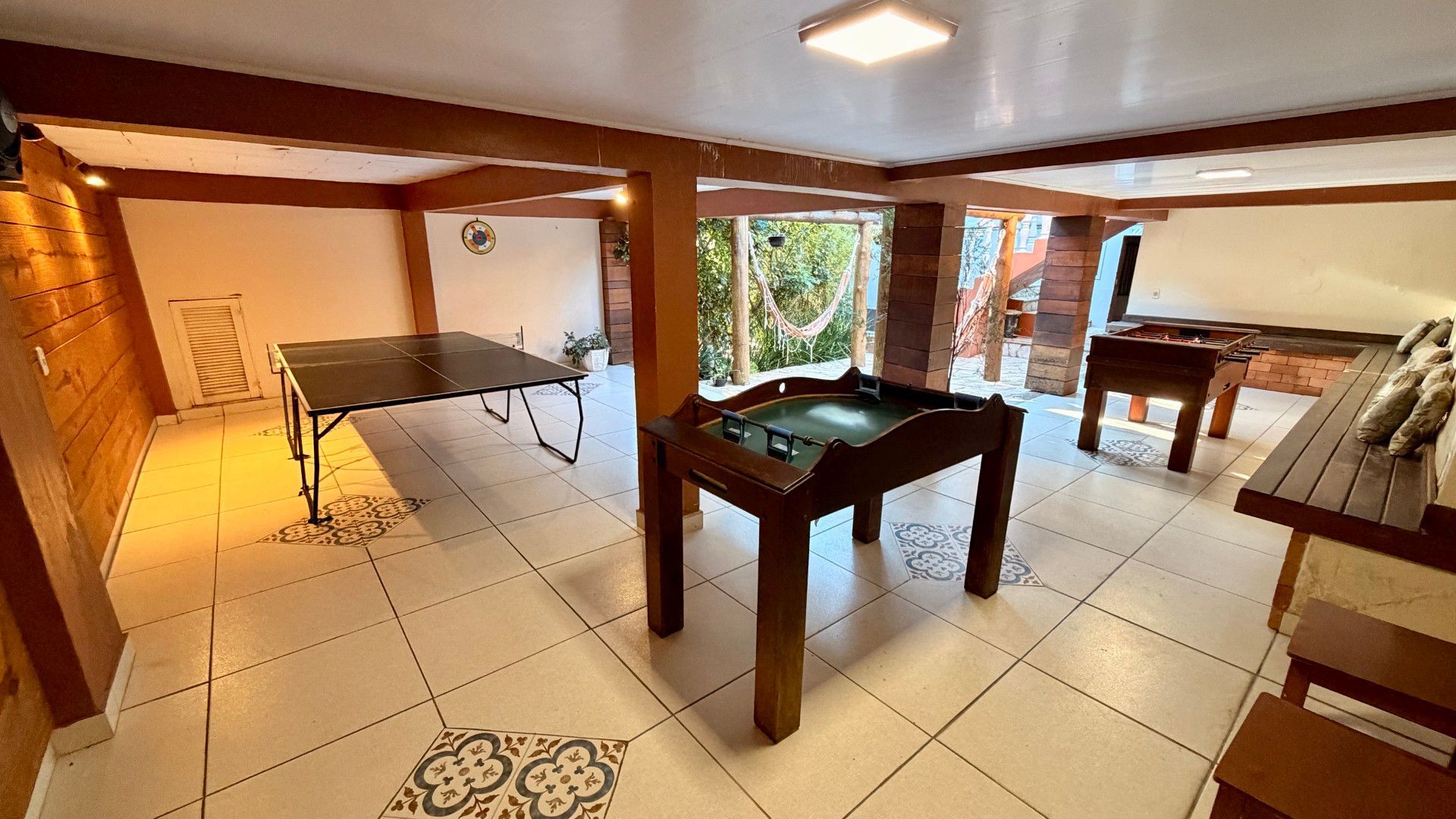 Sala ampla e iluminada, com piso de ladrilhos, mesa de bilhar e mesas de madeira, vista através de portas abertas e vigas