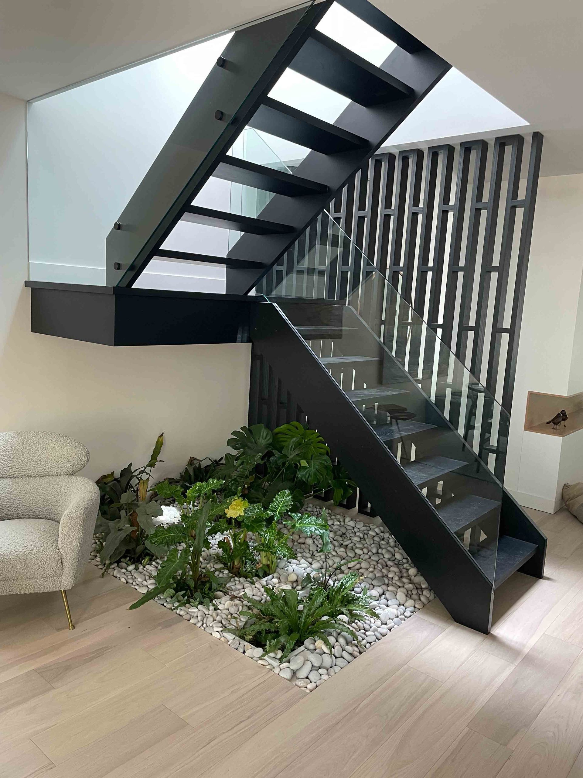 Escalier moderne en bois avec rampes et barreaux en métal noir, dans une maison lumineuse et aérée. L'entrée est carrelée et donne sur la salle à manger.