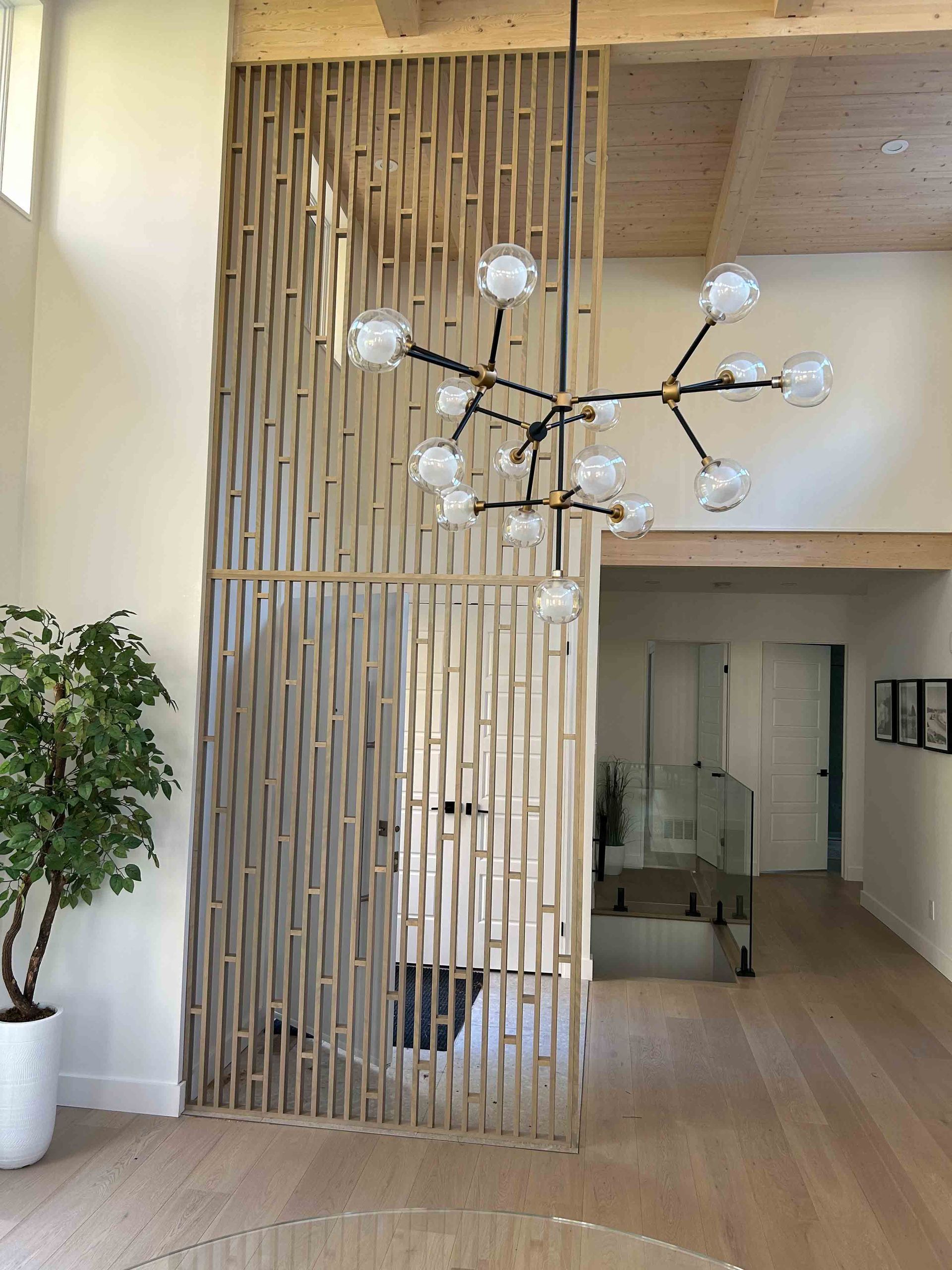 Intérieur moderne avec un lustre suspendu, une cloison verticale en lattes de bois, un arbre en pot et une porte vitrée.