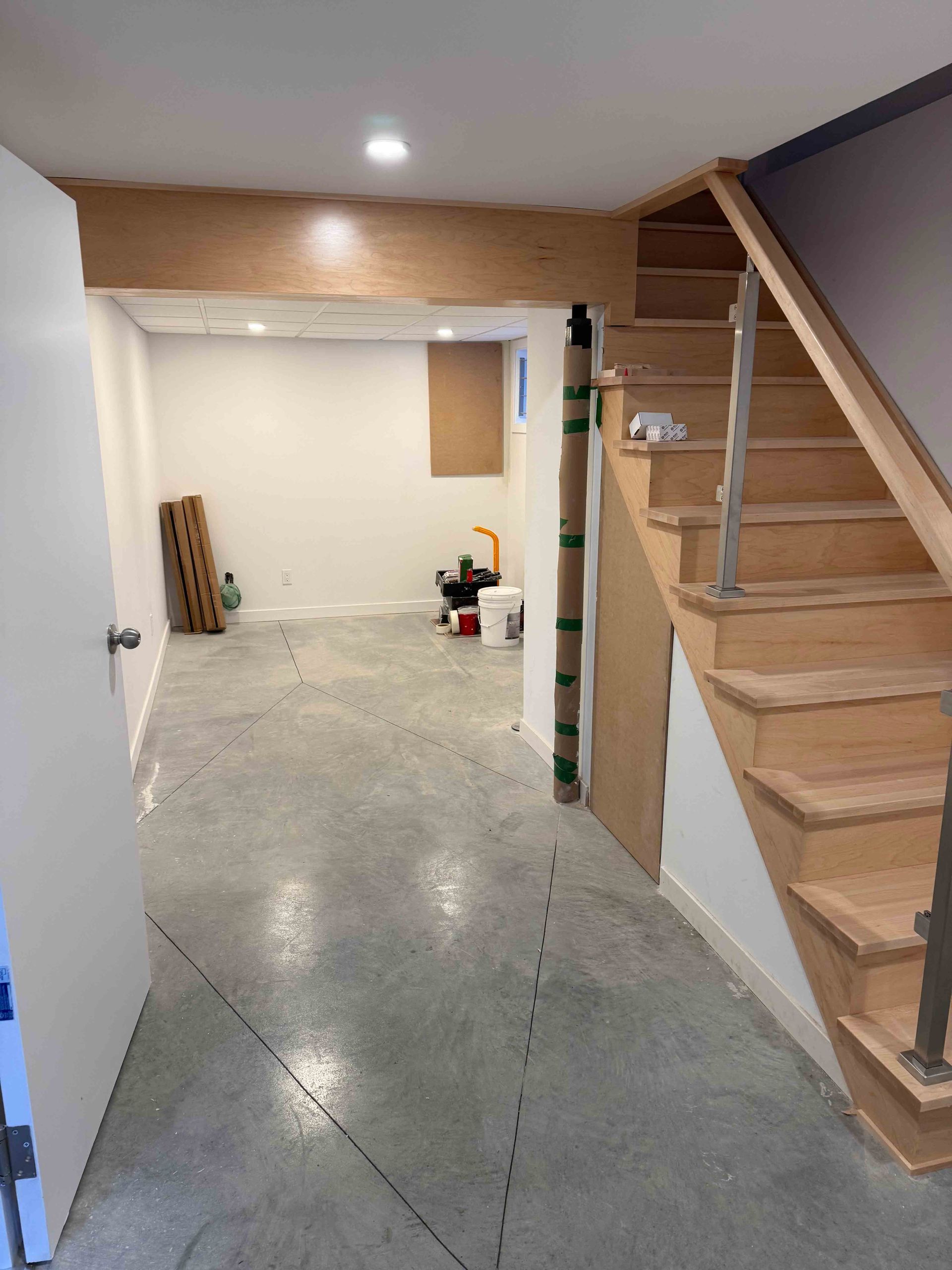 Pièce vide au sous-sol avec sol en béton brut et escalier en bois sur la droite.