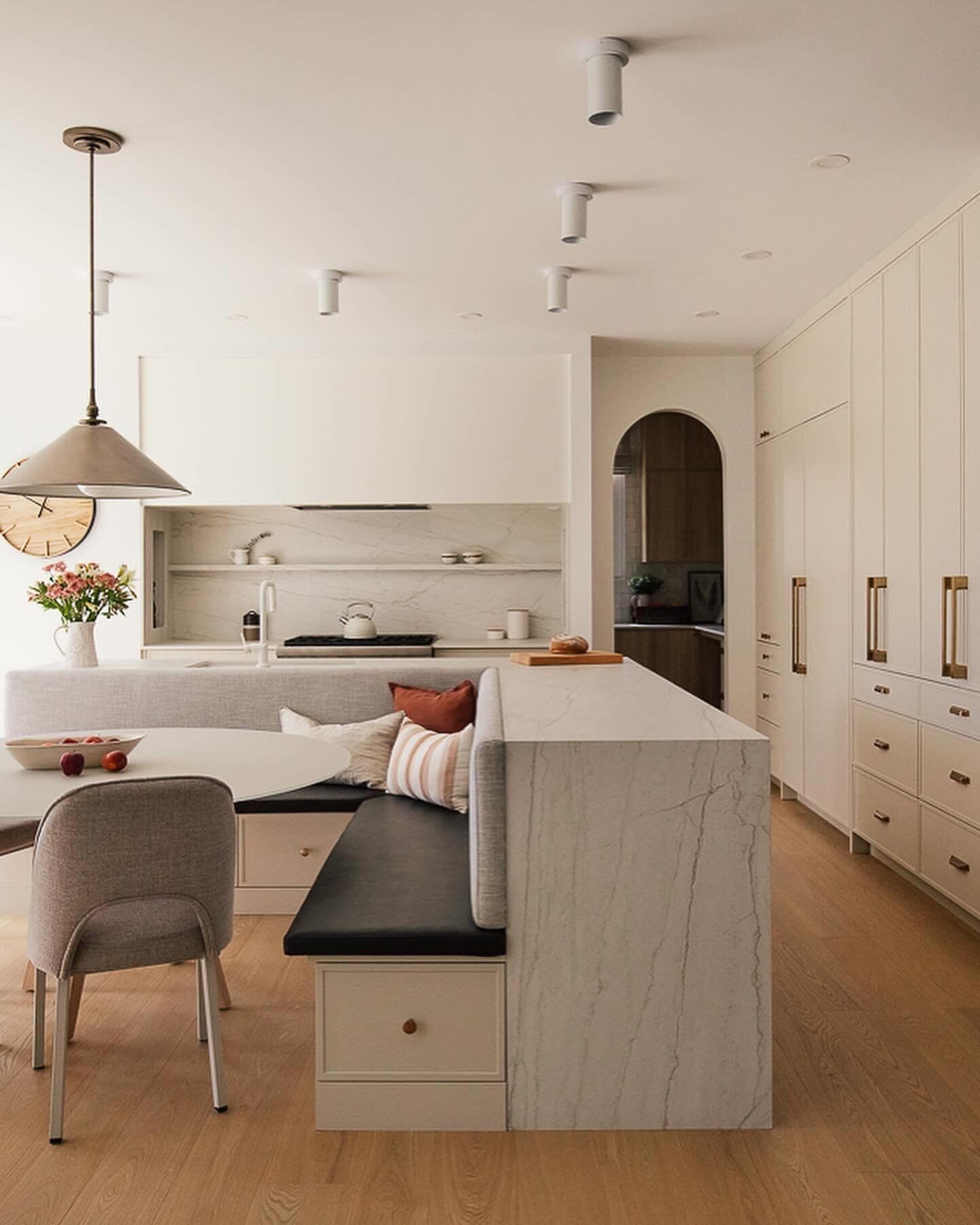 Cuisine moderne avec sièges intégrés, îlot en marbre et parquet clair. Elle présente des tons neutres, des accessoires dorés et un coin repas chaleureux.