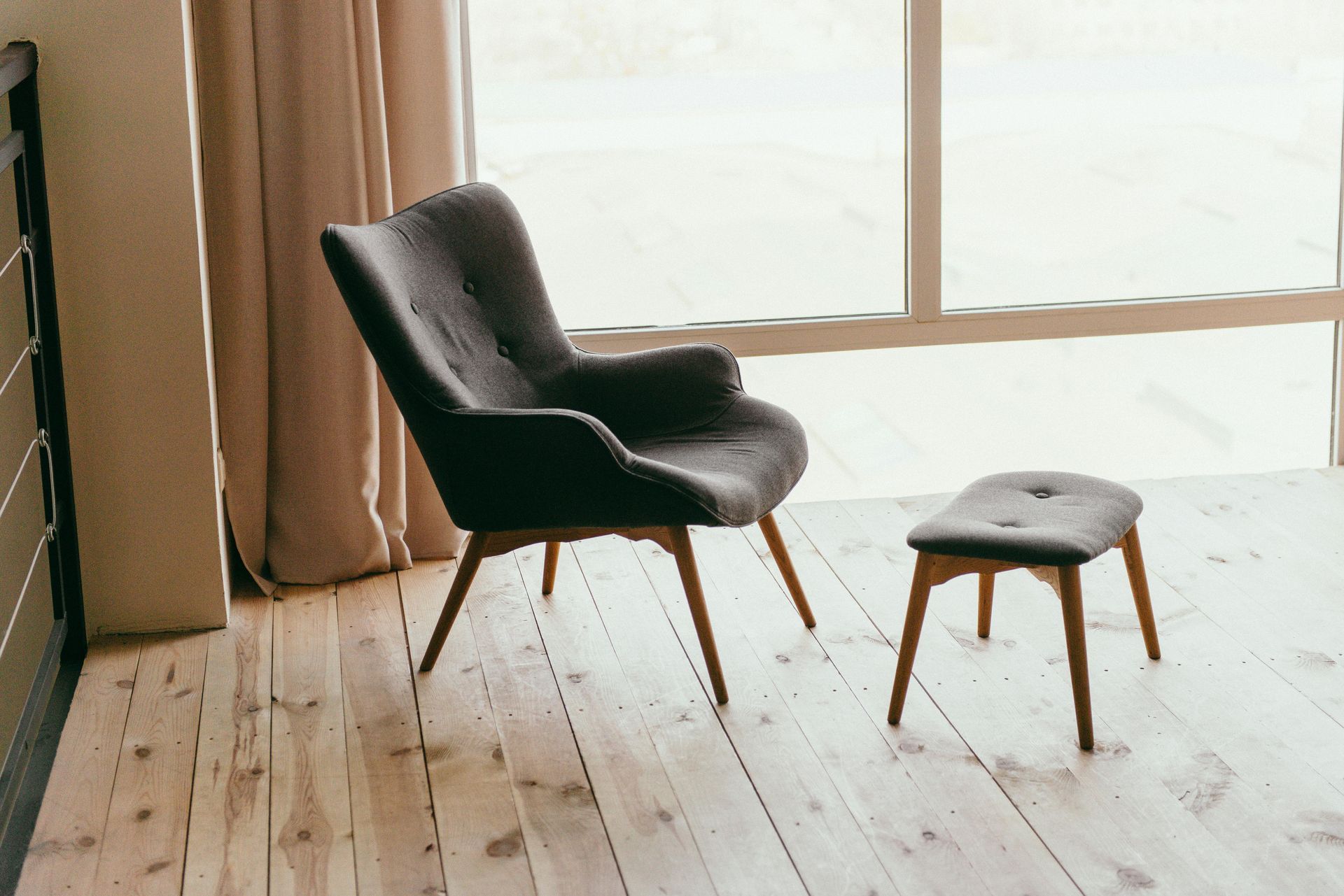Fauteuil et repose-pieds gris sur un parquet près d'une fenêtre aux rideaux transparents.