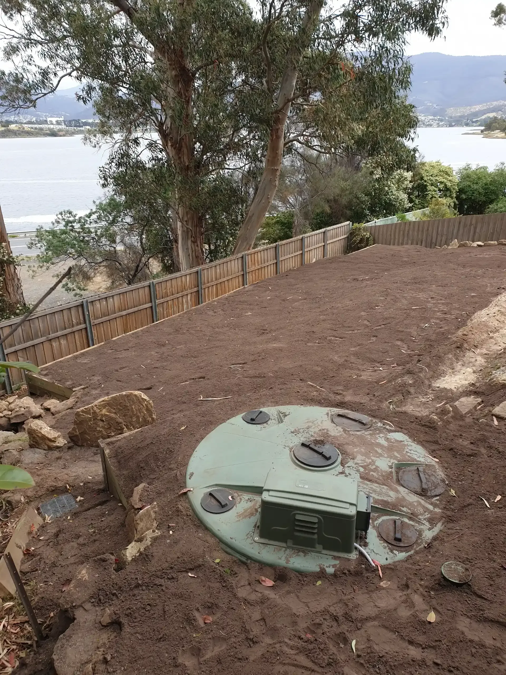 A green septic tank lid sits in a dirt-covered yard near a fence, overlooking a body of water and distant hills. — Lenny's Plumbing & Contracting in Granton, TAS