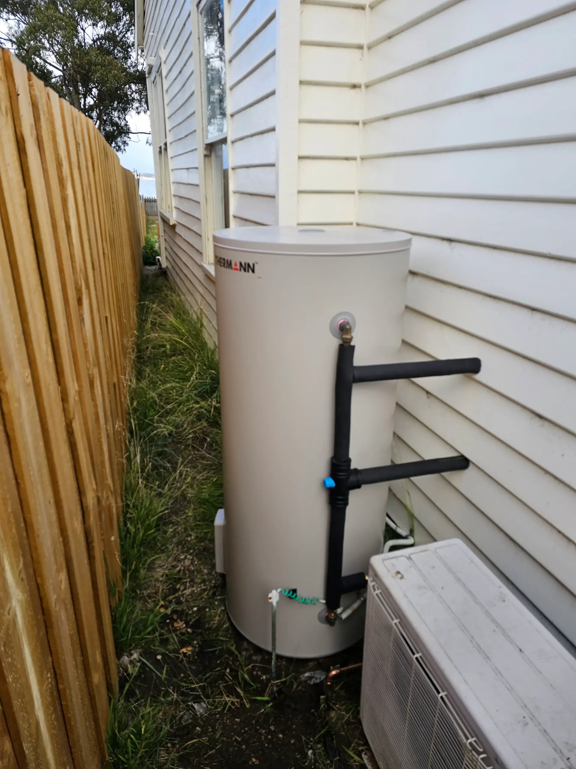 A tall, beige hot water heat pump tank positioned between a wooden fence and the white-sided wall of a house. — Lenny's Plumbing & Contracting in Granton, TAS
