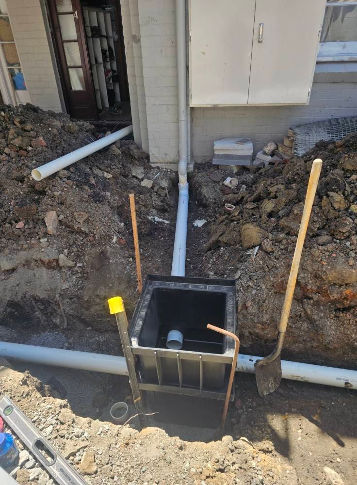 A storm water drain pit and PVC pipes installed in an outdoor trench beside a building, with two shovels nearby. — Lenny's Plumbing & Contracting in Granton, TAS