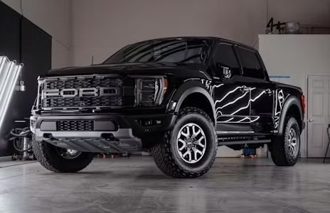 Black Ford F-150 Raptor pickup truck in a well-lit garage, glossy finish, large off-road tires.
