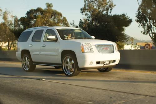 A white GMC Yukon SUV driving on a highway with custom chrome wheels.