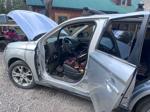 Silver car with hood and driver door open, showing the interior and engine area outside a building