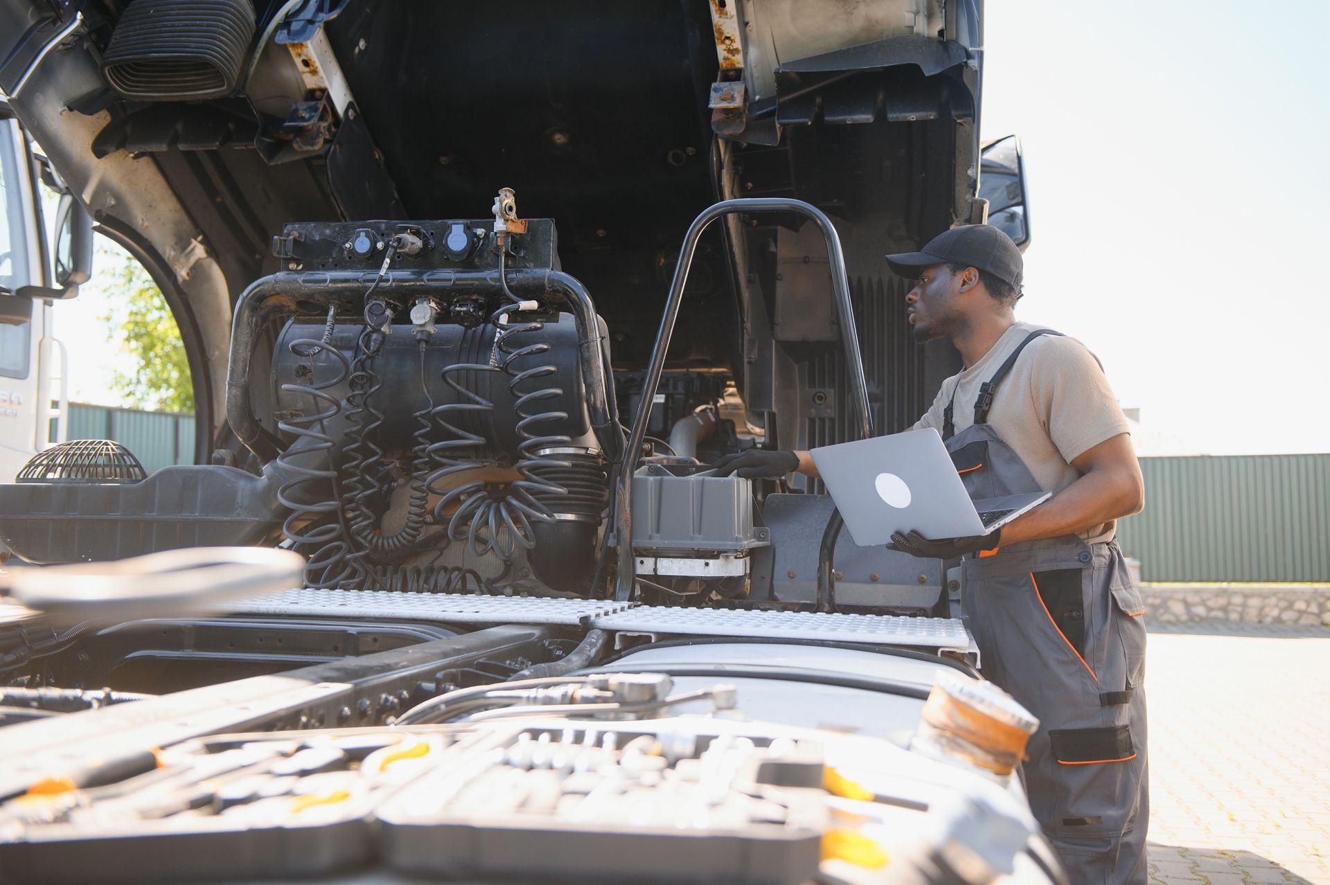 Mechanic using a laptop beside an open truck engine in a sunlit garage
