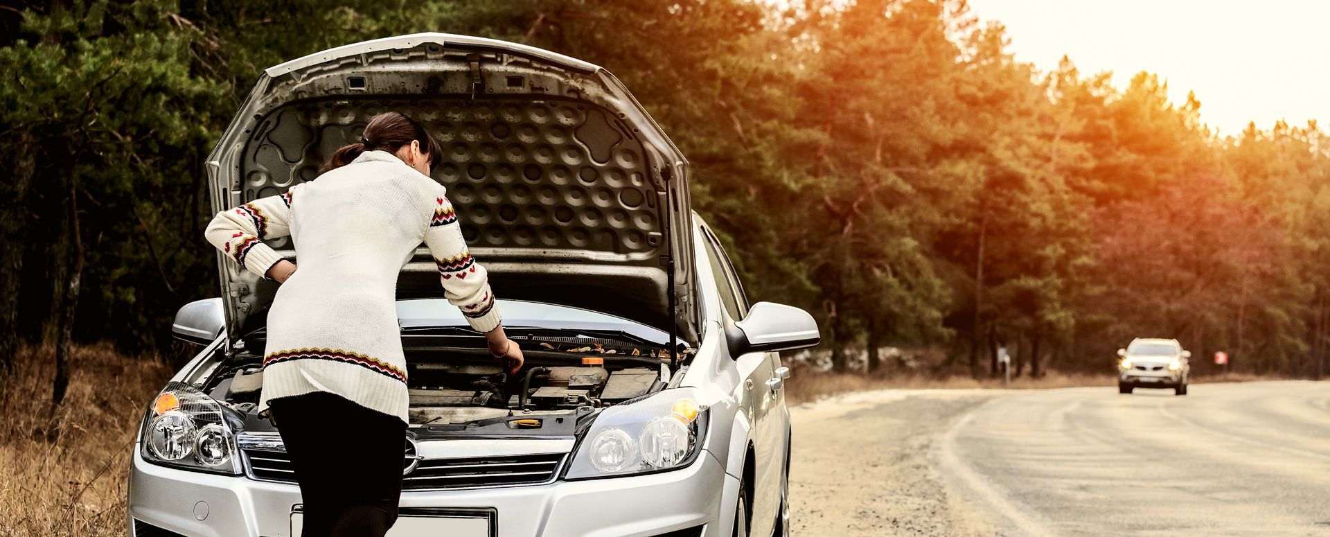 Person checks under the hood of a silver car on a roadside with autumn trees nearby