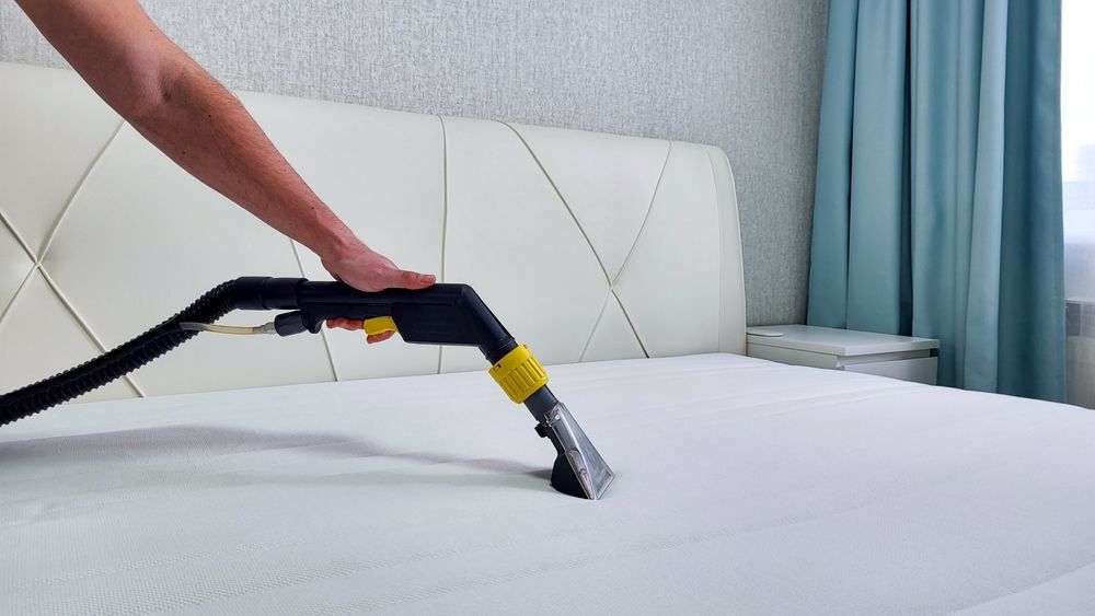 A person vacuuming a white bed mattress with a handheld upholstery tool in a bedroom