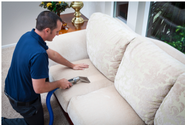 Person vacuuming a gray sofa with a handheld vacuum, wearing gloves and a pink shirt.