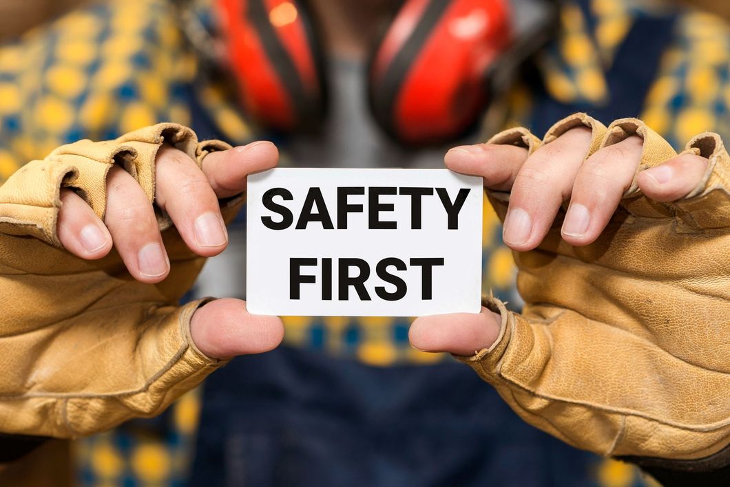Hands in work gloves holding a “SAFETY FIRST” sign in front of safety gear.