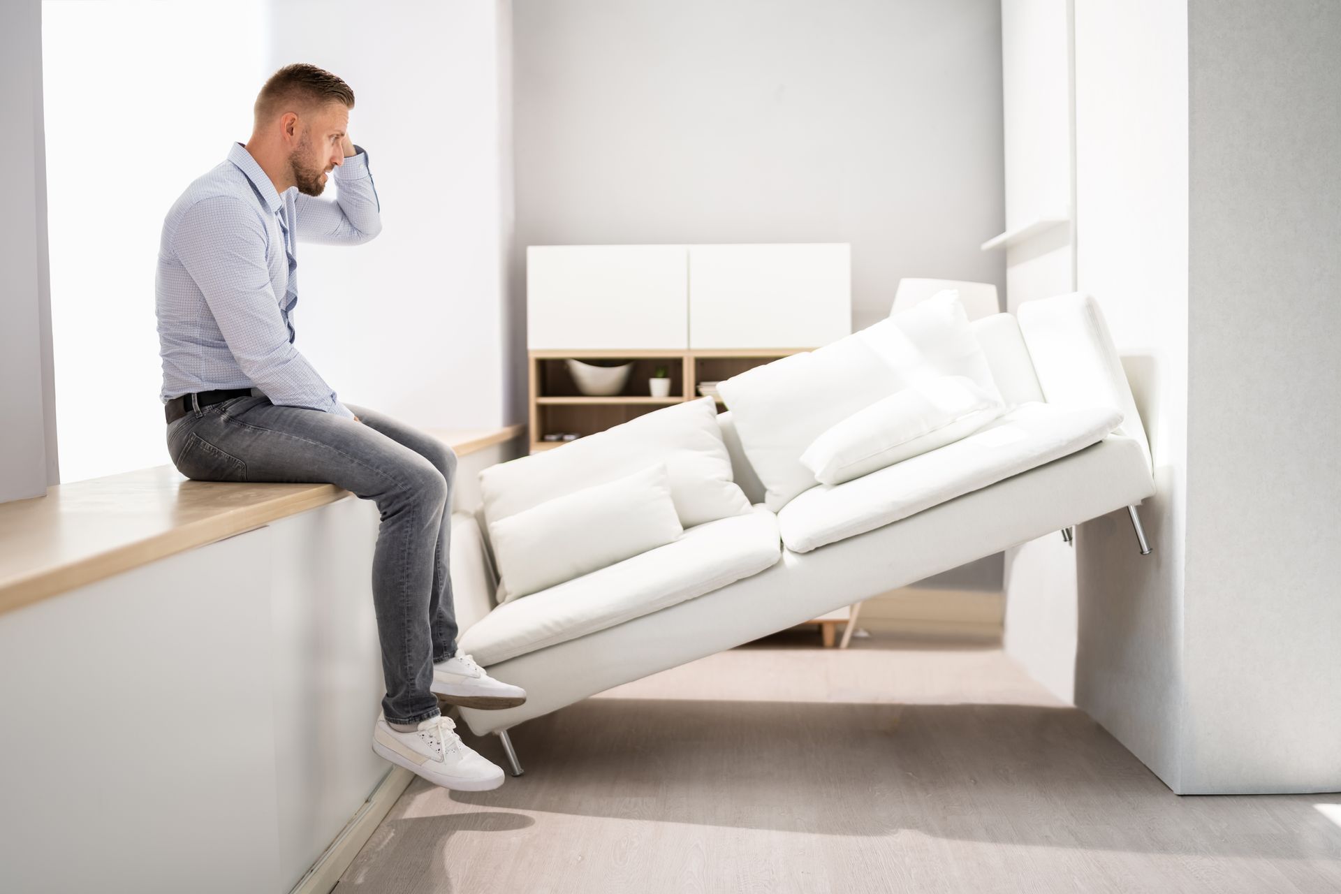 Man sitting by a window, surrounded by all white furniture and a white sofa stuck between walls.
