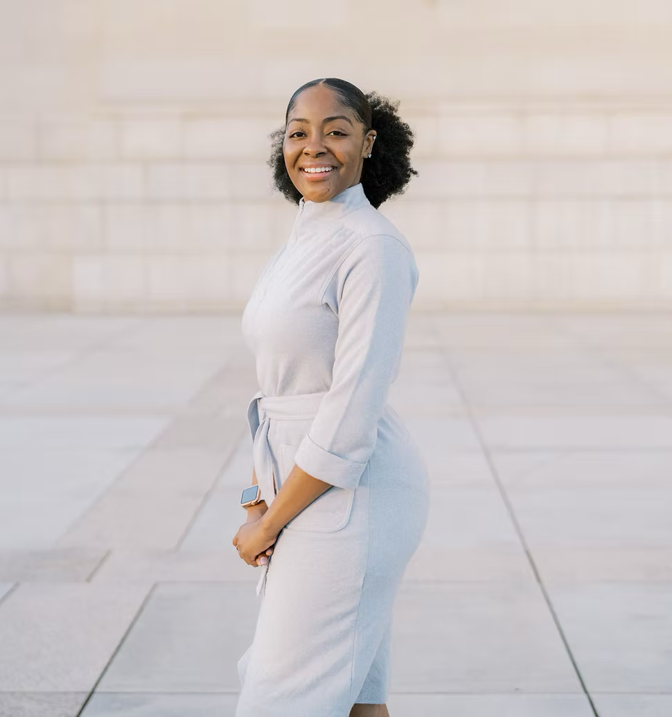 A person stands sideways, smiling at the camera, wearing a light grey belted jumpsuit against a stone-tiled background.