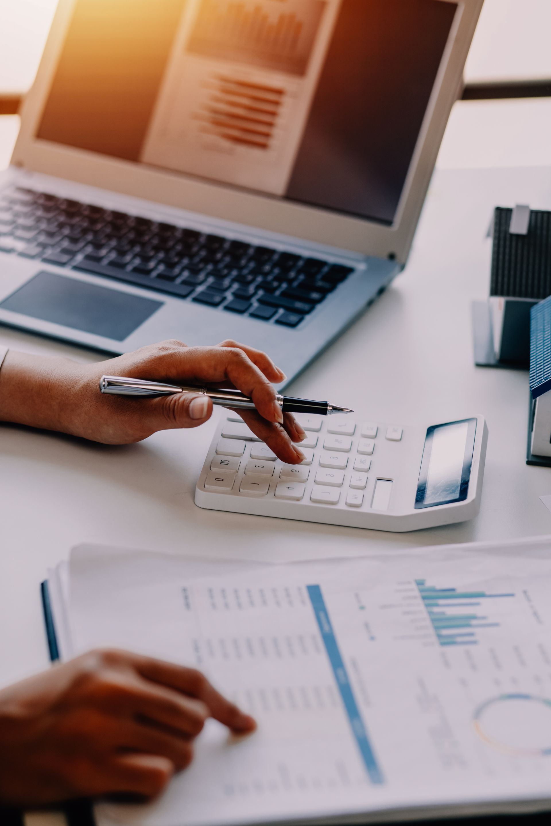 A person uses a white calculator and reviews financial charts at a desk with a laptop.