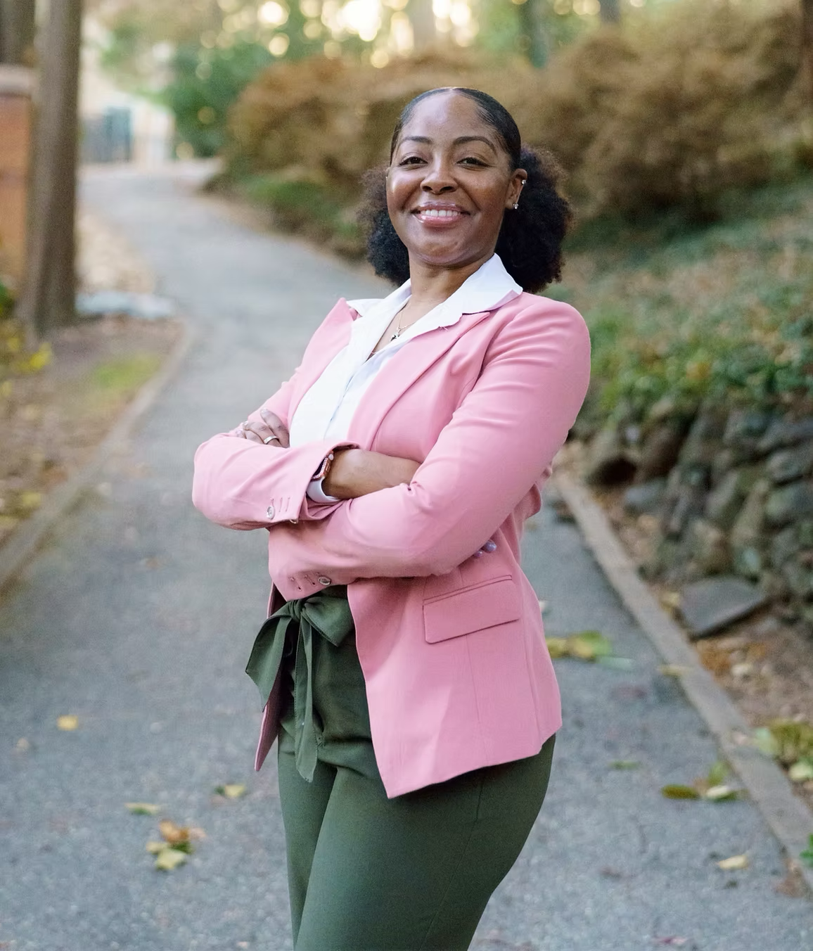 A person in a pink blazer and olive green trousers stands with arms crossed on a paved path outdoors.