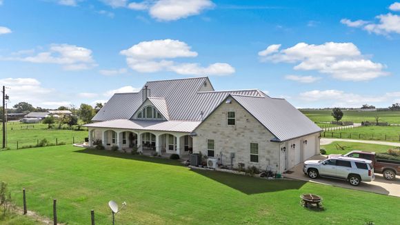 A large white house with a gray roof is sitting on top of a lush green field.