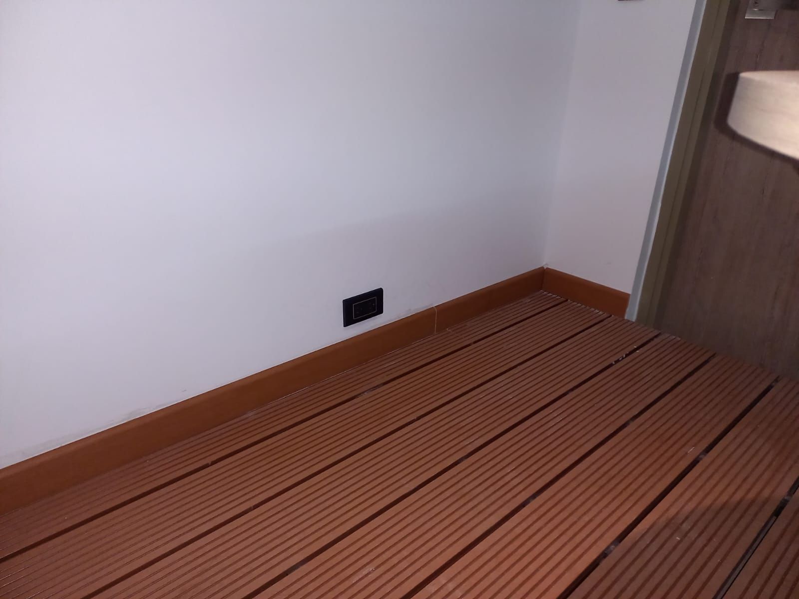 Rincón vacío con pared blanca, suelo de madera marrón y un enchufe de pared negro.
