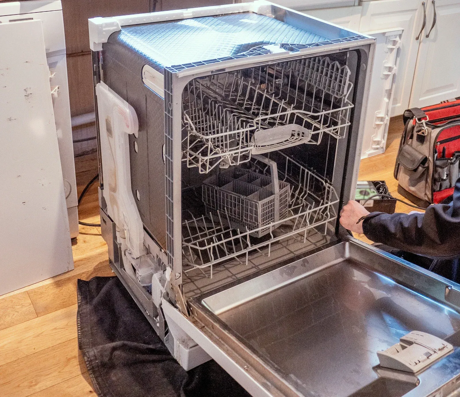 Open dishwasher with rack and utensils beside a stainless steel sink on a kitchen floor