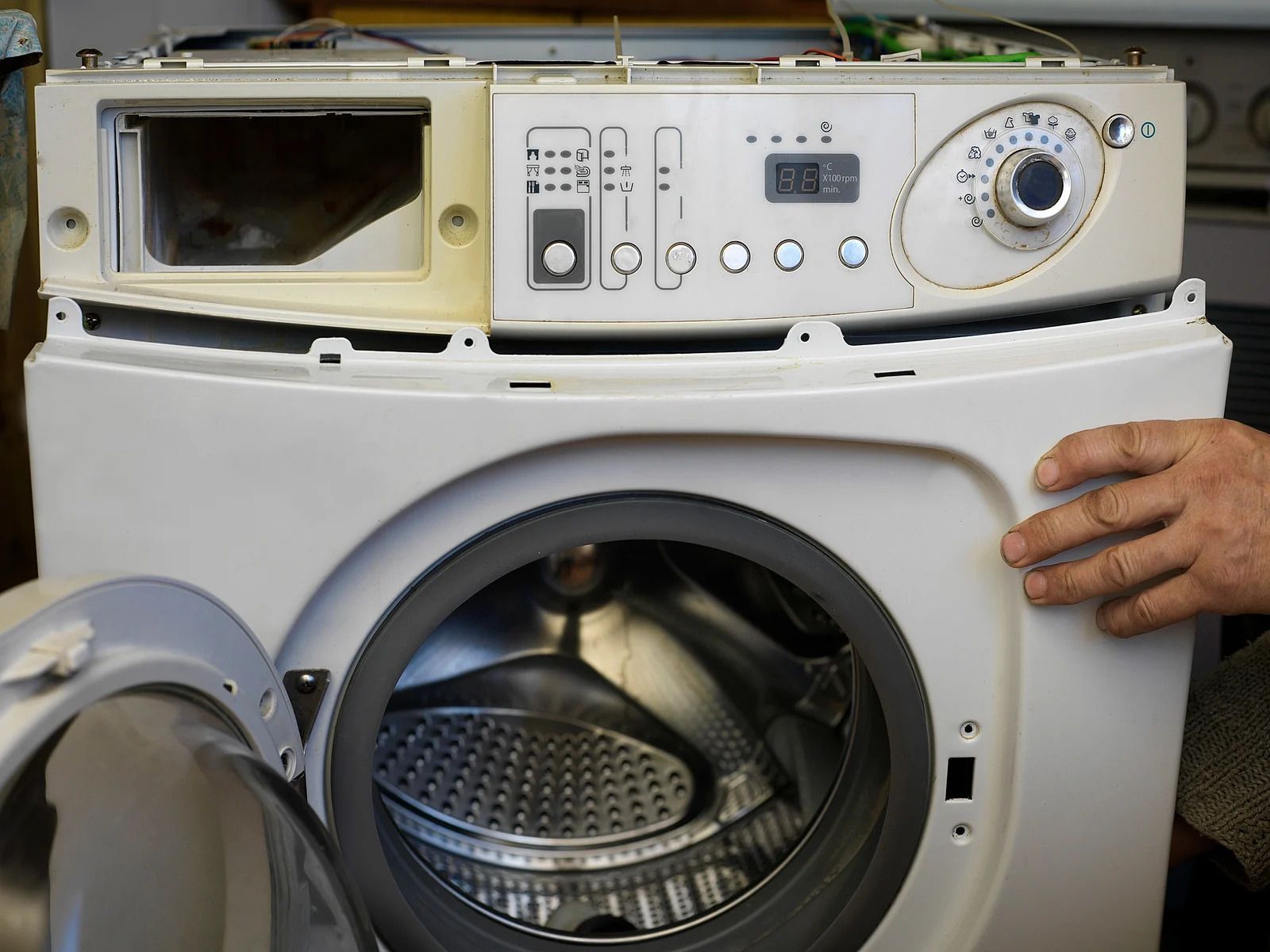 Partially disassembled white front-loading washing machine with control panel exposed and a hand holding the side