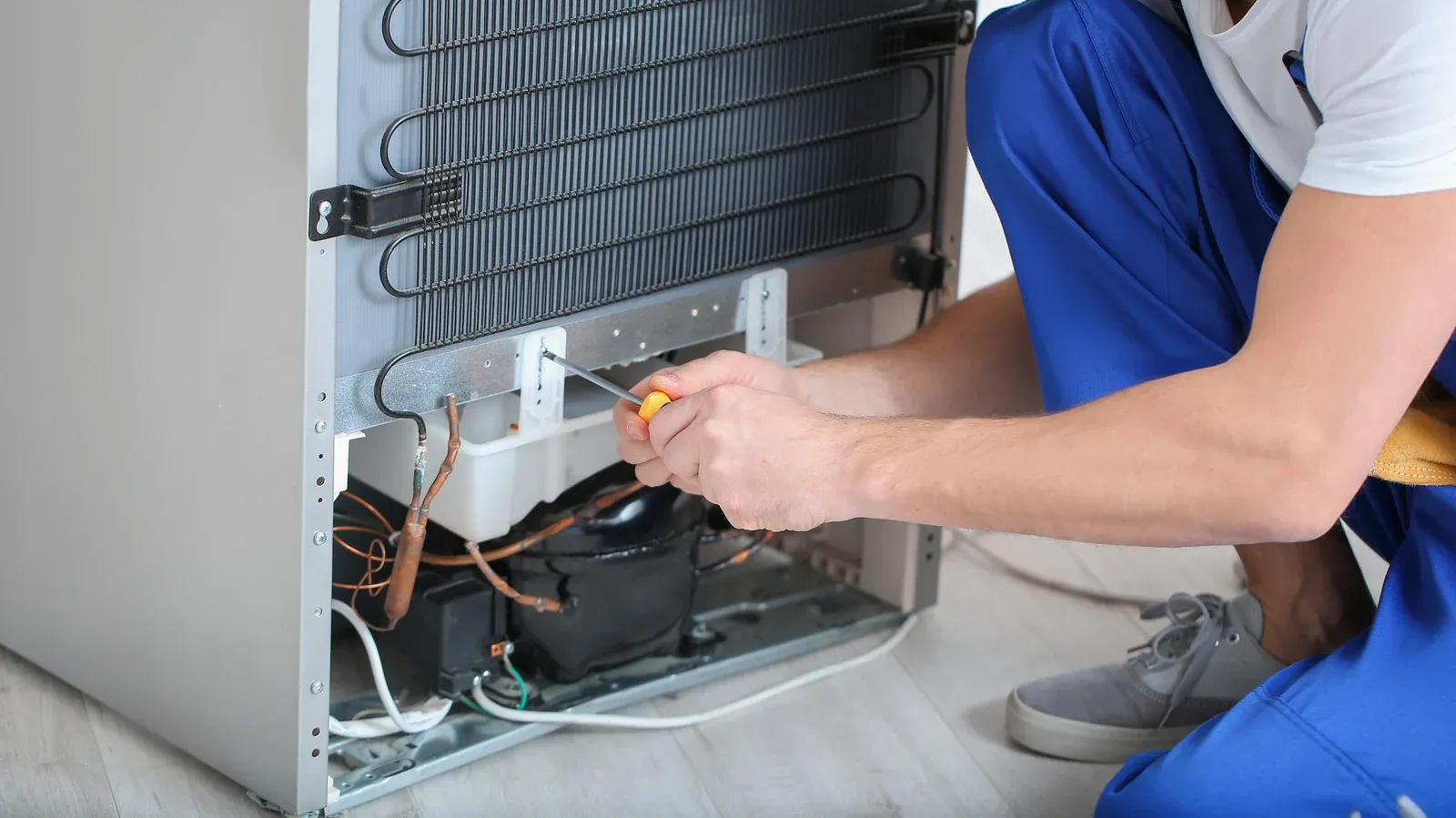 Technician repairing a refrigerator, working on exposed wiring and compressor behind the open back panel.