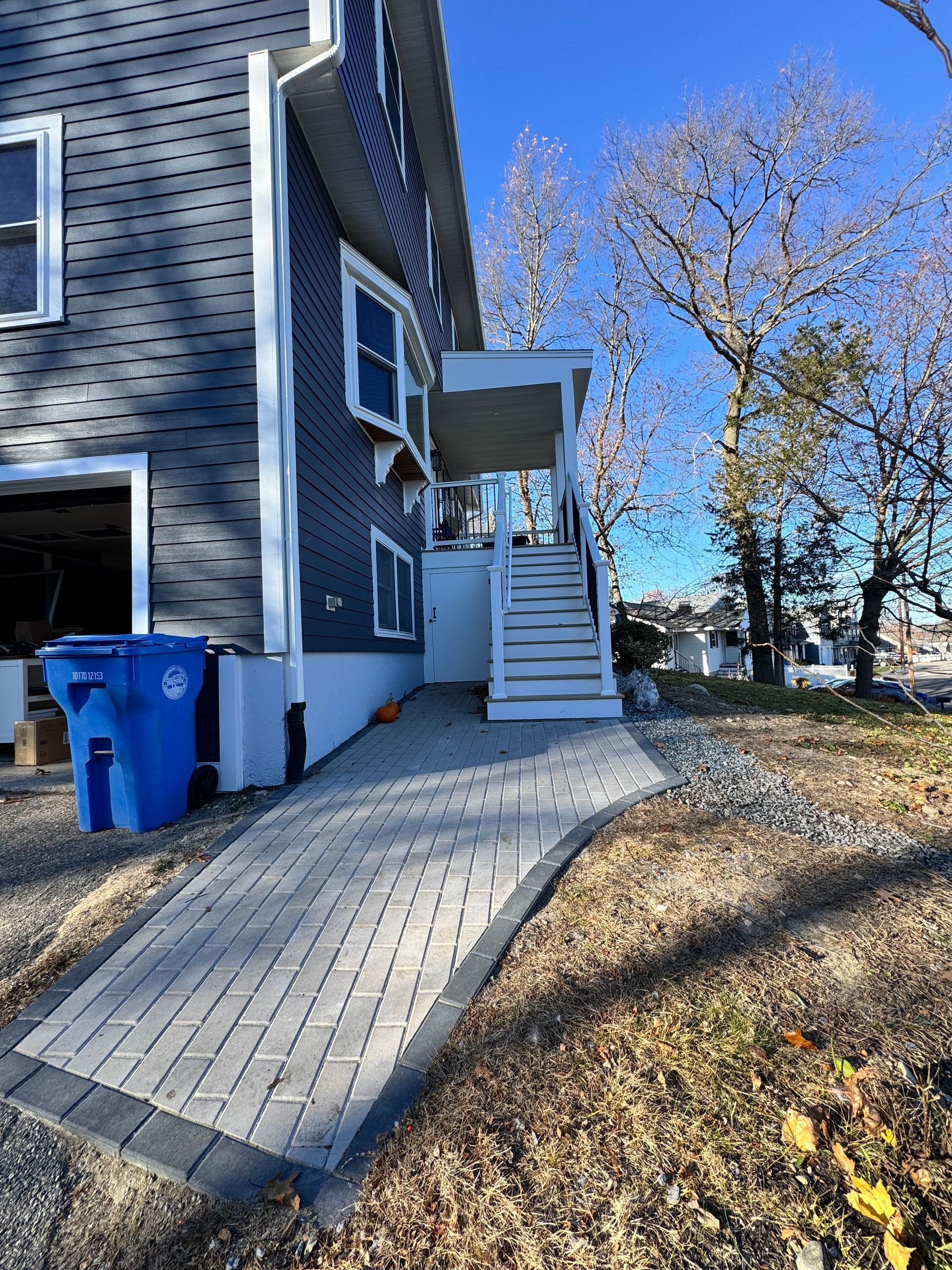 Brick pathway leading to a building with stairs and a small deck. A blue trash bin sits on the side.