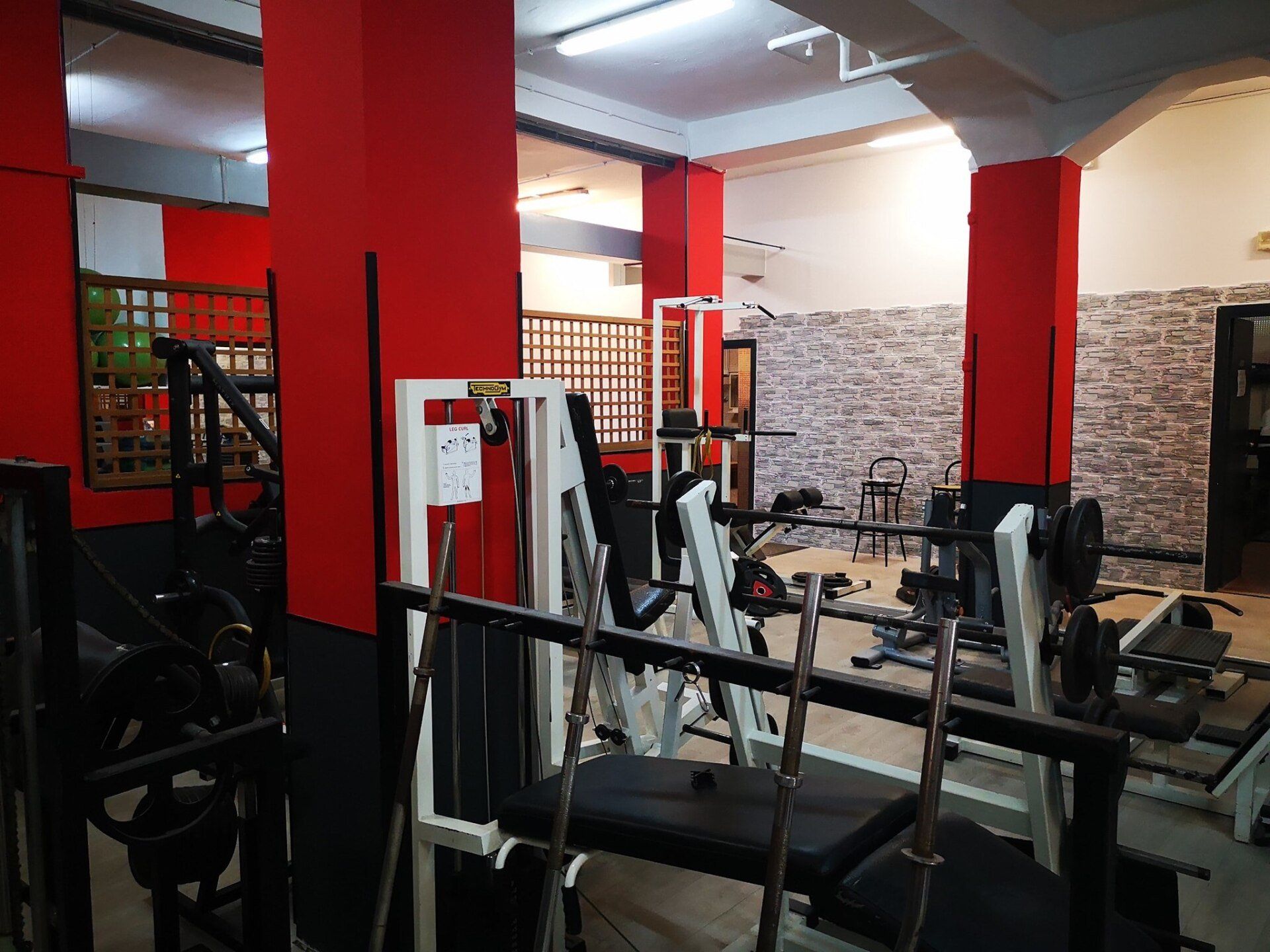 Interno di una palestra con attrezzature per il sollevamento pesi. Pareti rosse e grigie con vari macchinari.