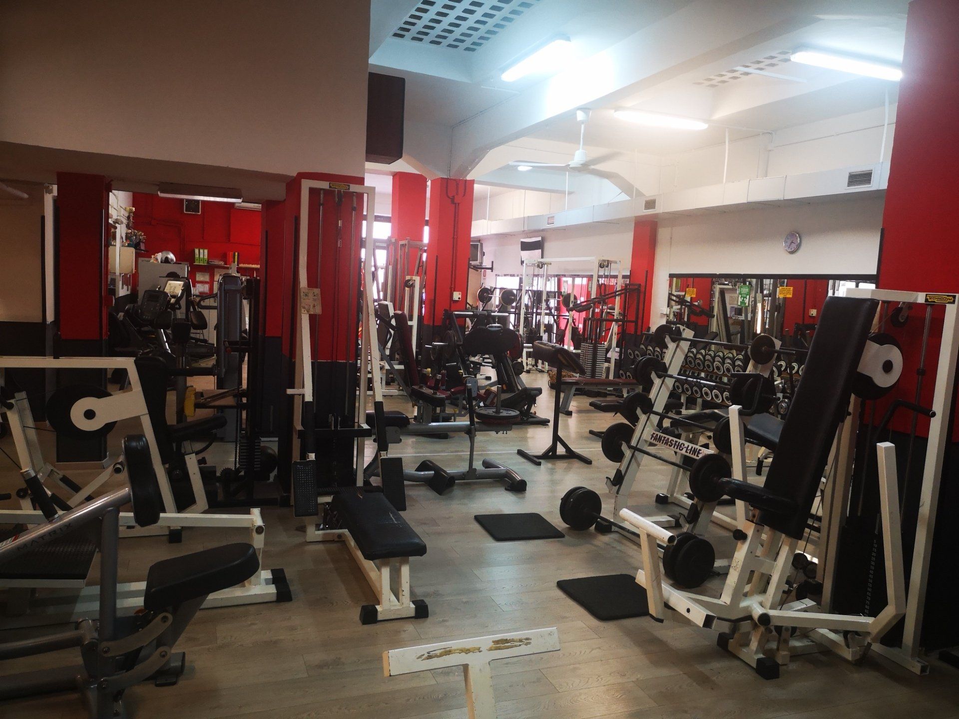 Interno della palestra con attrezzi per pesi e panche, pareti rosse e bianche, illuminazione intensa.