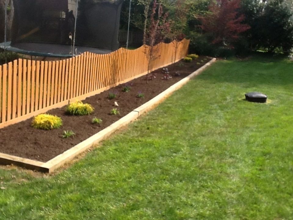 Wooden fence along a landscaped flower bed with yellow plants; a trampoline is visible in the background.