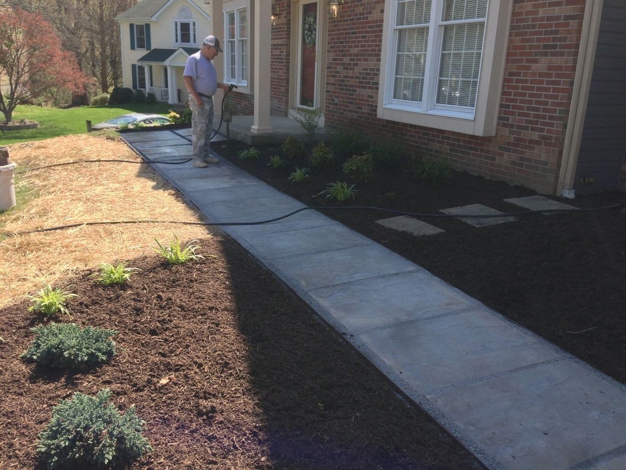 Man sprays a concrete walkway in front of a brick house with new landscaping.