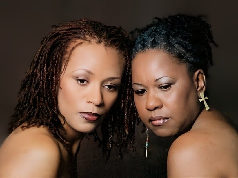 Two people with locs positioned closely together against a dark background, both wearing contemplative expressions.