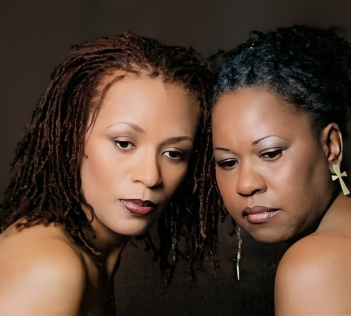Two people with locs posing closely together against a dark background, both looking down with neutral expressions.