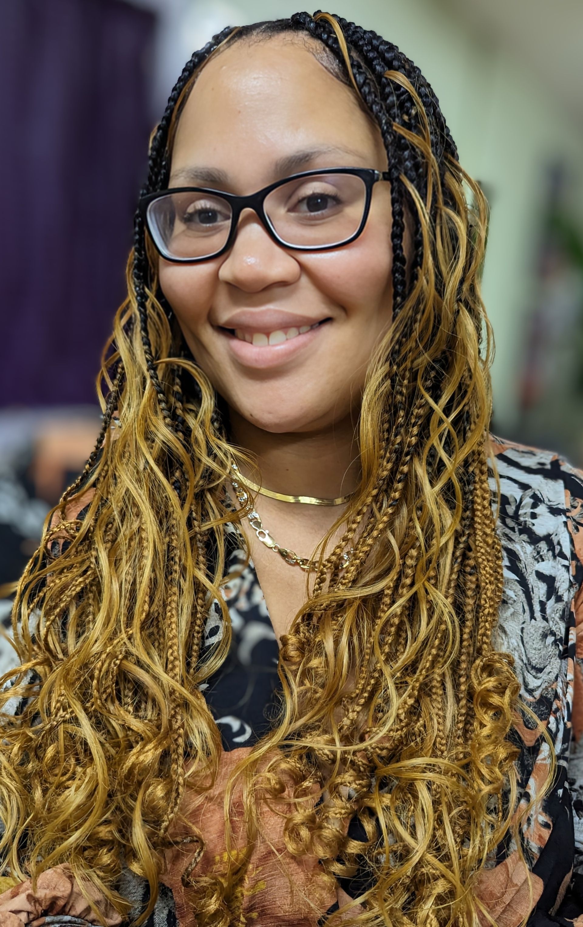 A smiling person wearing glasses and long, wavy, golden-brown braided hair, wearing a patterned top and gold necklaces.