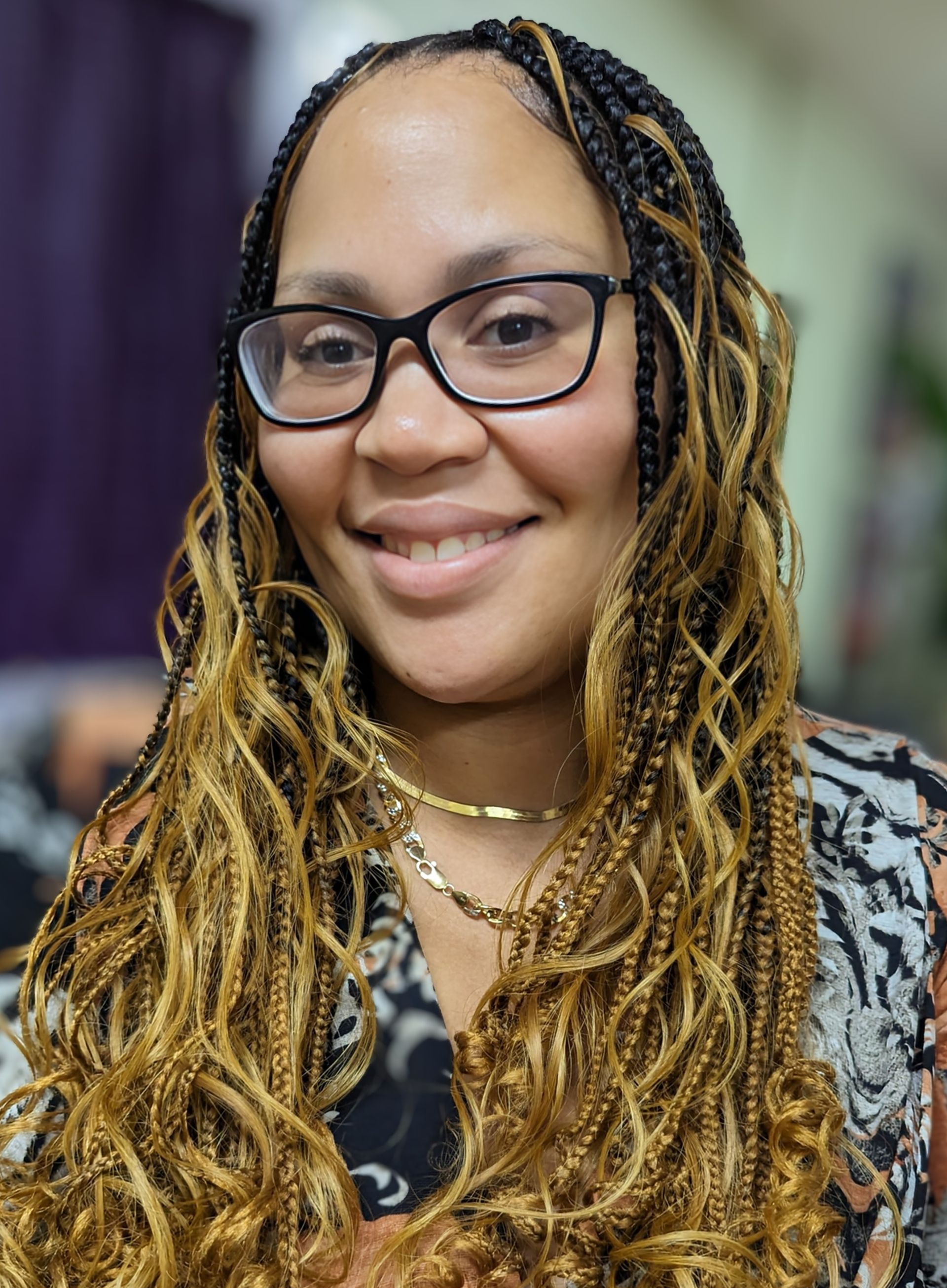 A smiling person with long, blonde-highlighted braided hair wearing glasses and a gold chain necklace.