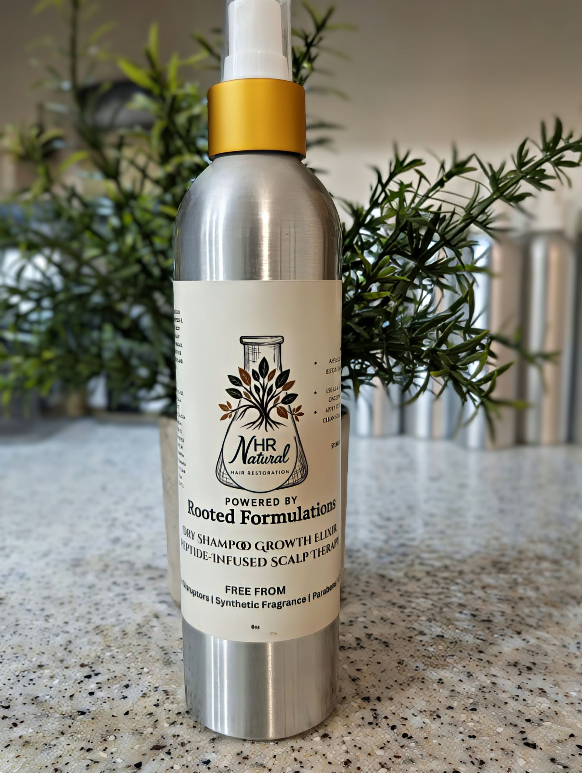 A silver spray bottle with a gold cap and a cream label featuring a tree icon, sitting on a countertop with green plants.