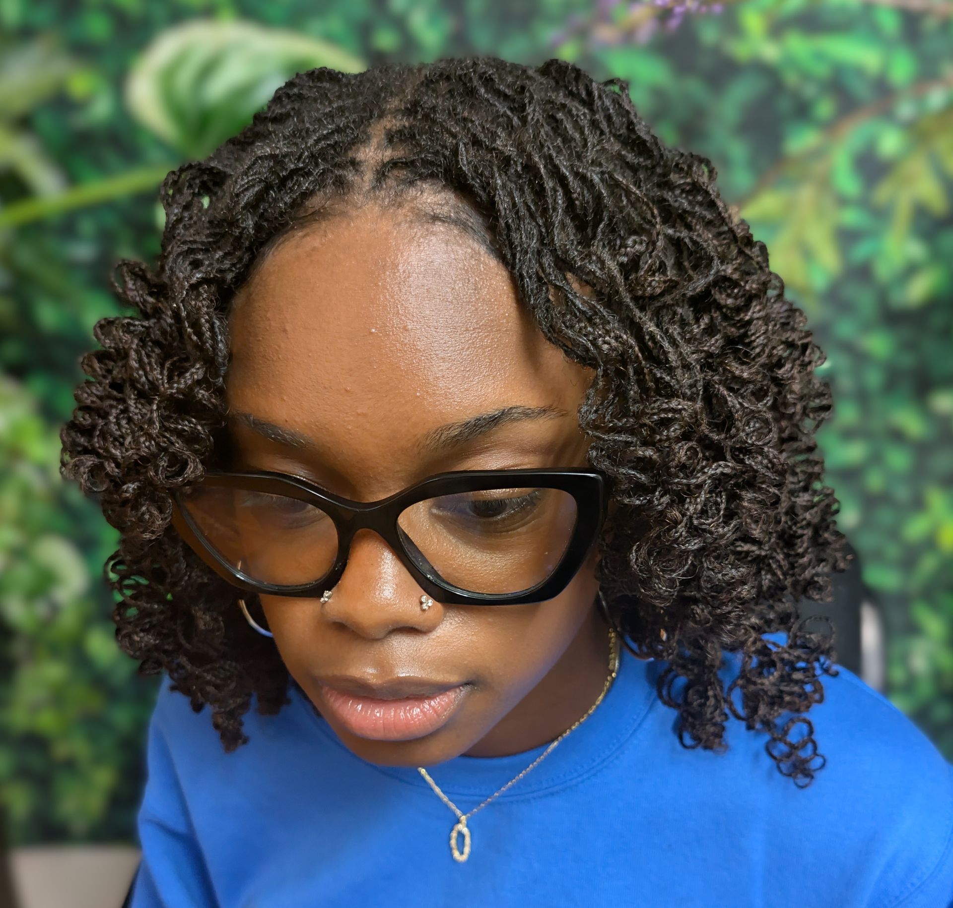 A person with shoulder-length curly dark locs wears black cat-eye glasses and a blue shirt against a green leafy background.