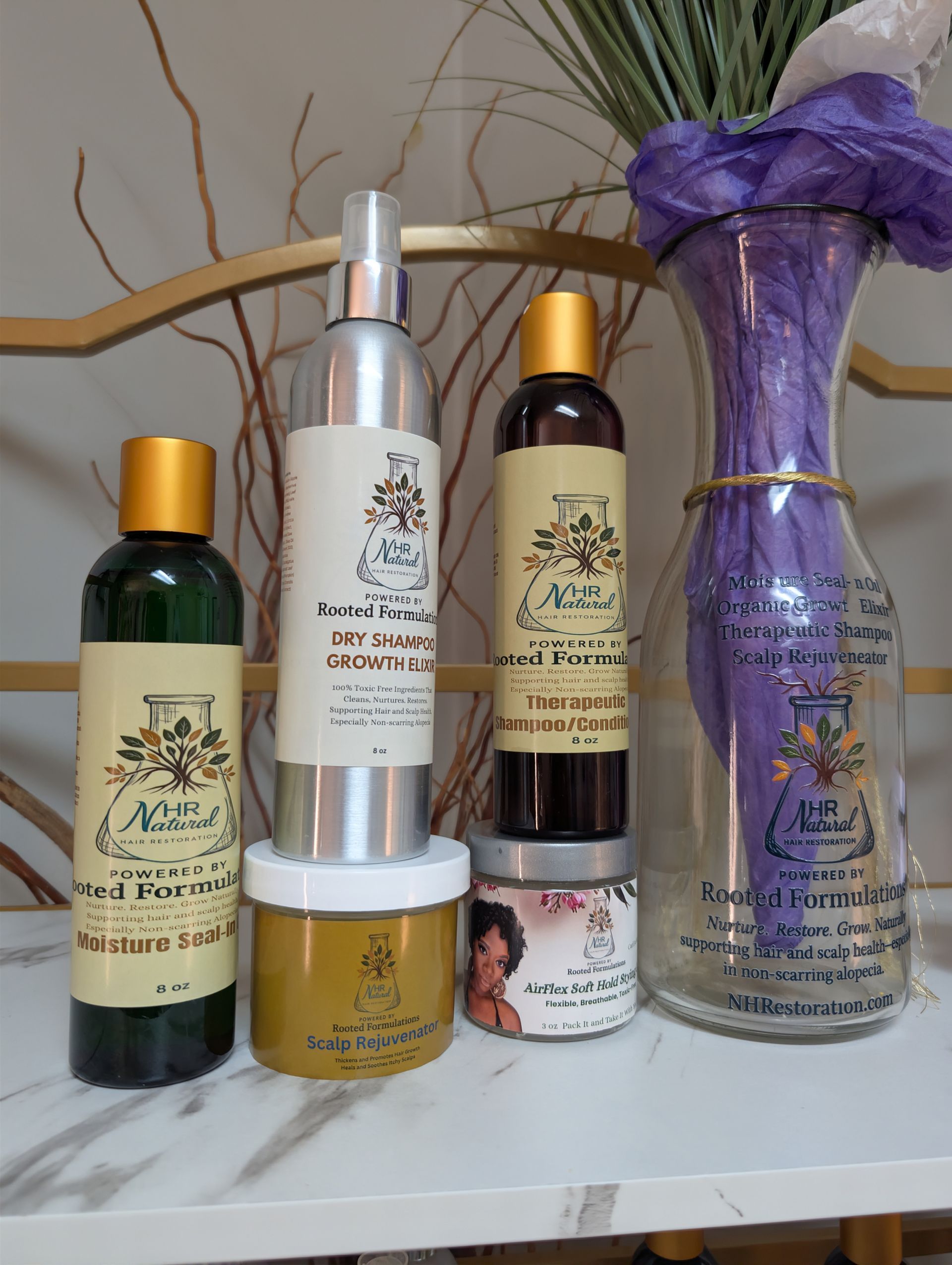 Four bottles and two jars of hair care products arranged on a white shelf next to a glass vase with purple fabric inside.