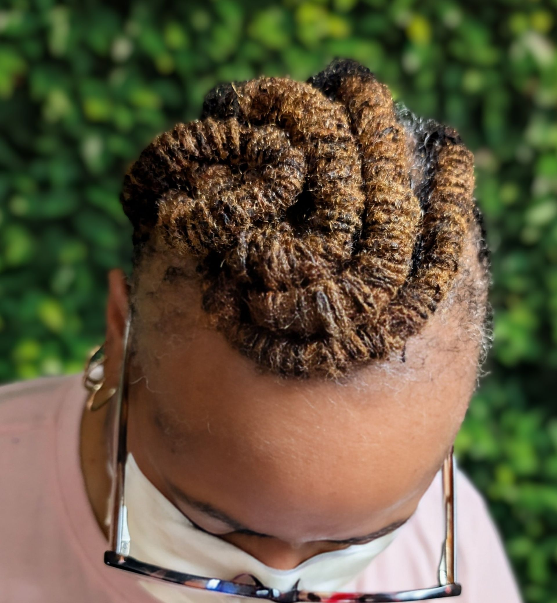 Brown locs styled in a tight spiral bun atop the head, worn by a person wearing glasses and a light-colored top.