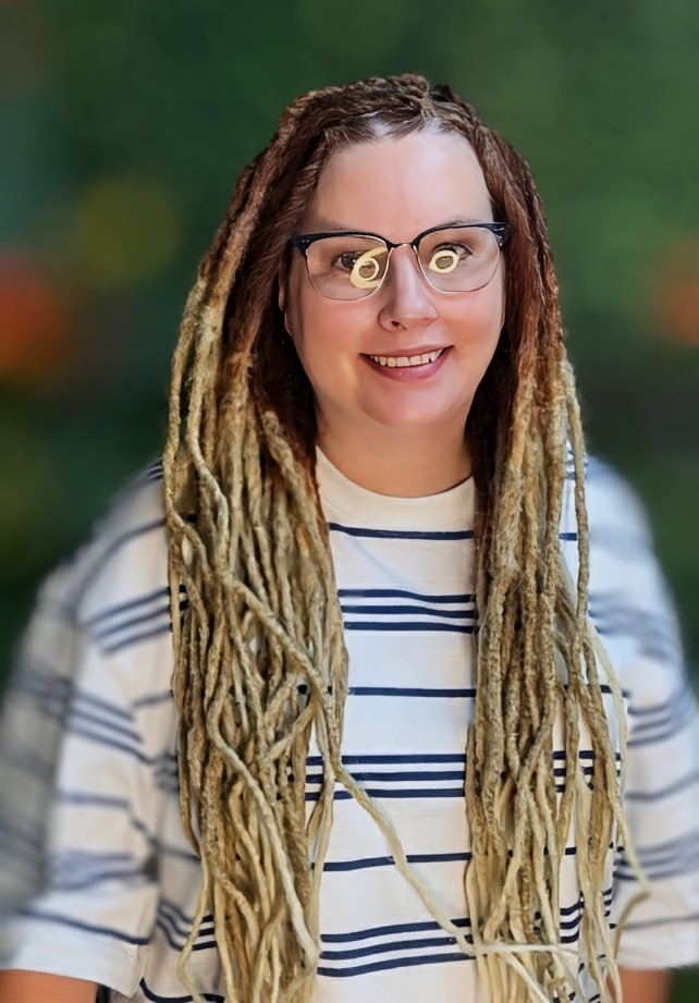 A smiling person with long, blonde dreadlocks wears glasses and a white and blue striped t-shirt against a blurred backdrop.
