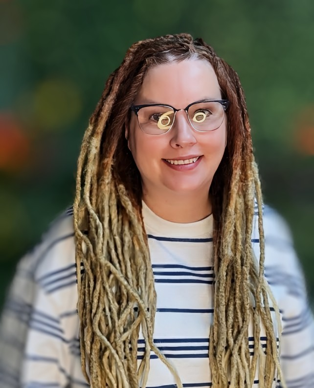 A person with long, blonde dreadlocks and glasses smiles, wearing a blue and white striped shirt against a blurred background.