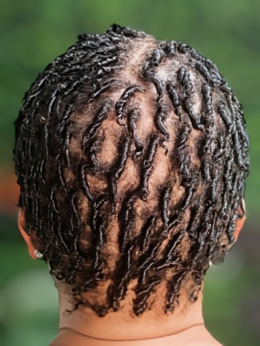 Back view of short hair styled in thin, defined, dark-colored finger coils against a blurred green background.